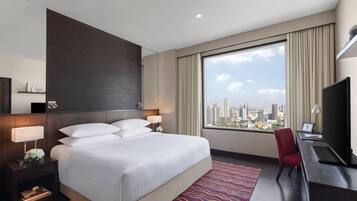 A hotel room with a large bed, a desk with a TV, and a view of a cityscape through a large window.