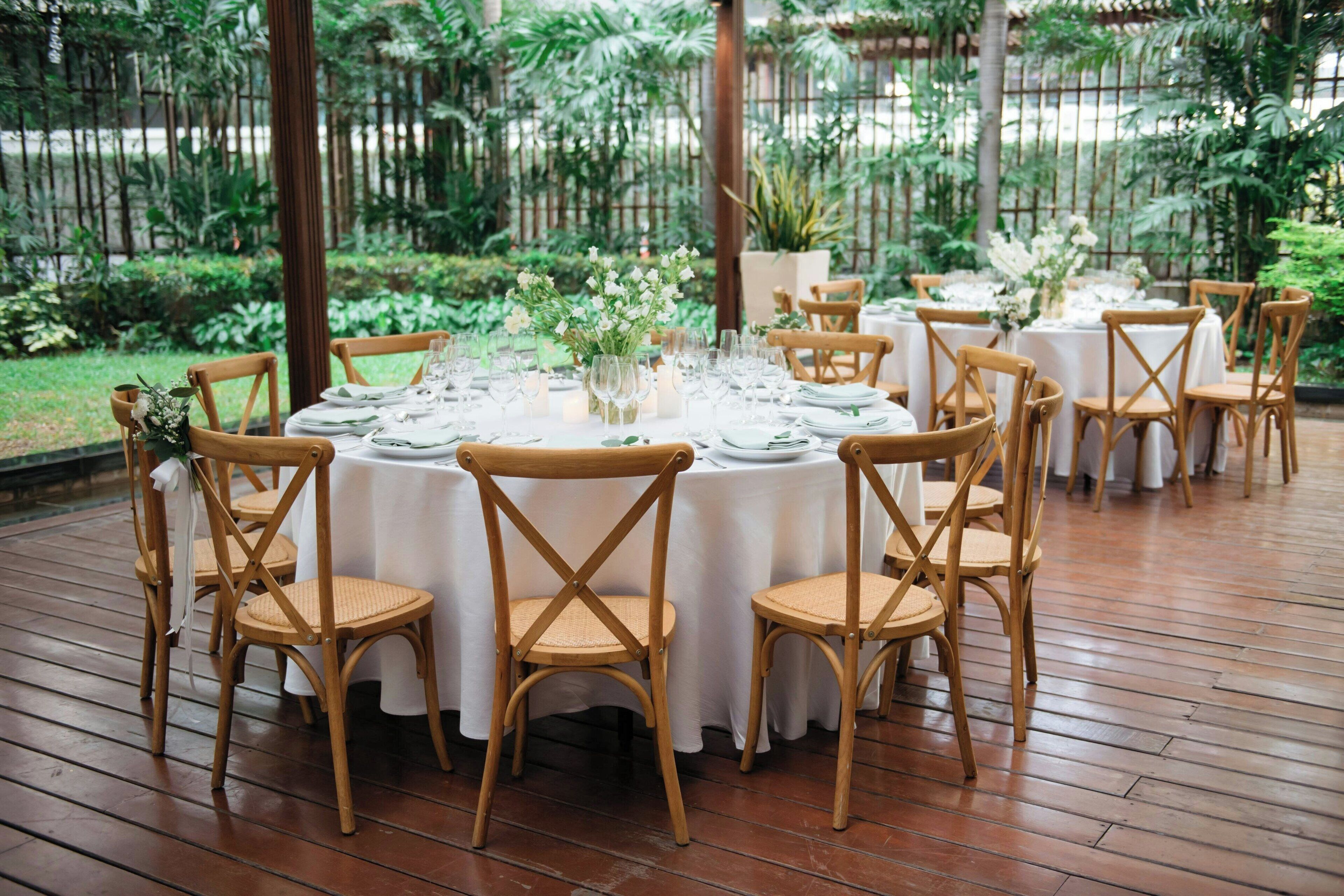 A round table set with white tablecloths, wooden chairs, and floral centerpieces in an outdoor garden setting.