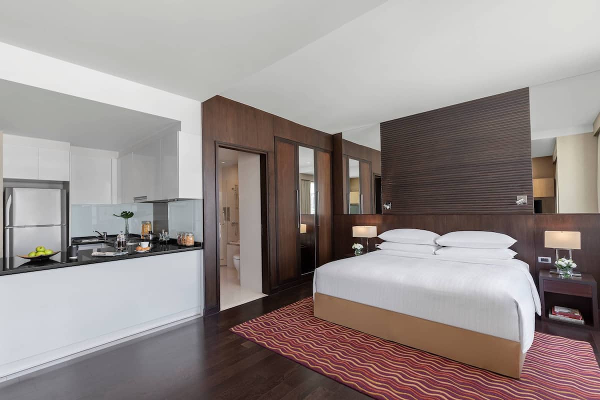 A modern hotel room with a large bed, a small bedside table with a lamp, a built-in wardrobe, and a kitchenette with a sink and countertop.