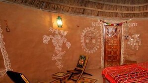 Une chambre traditionnelle avec un plafond de chaume, un lit recouvert d’une courtepointe à motifs rouges, une petite table avec une chaise et un mur orné de motifs décoratifs.