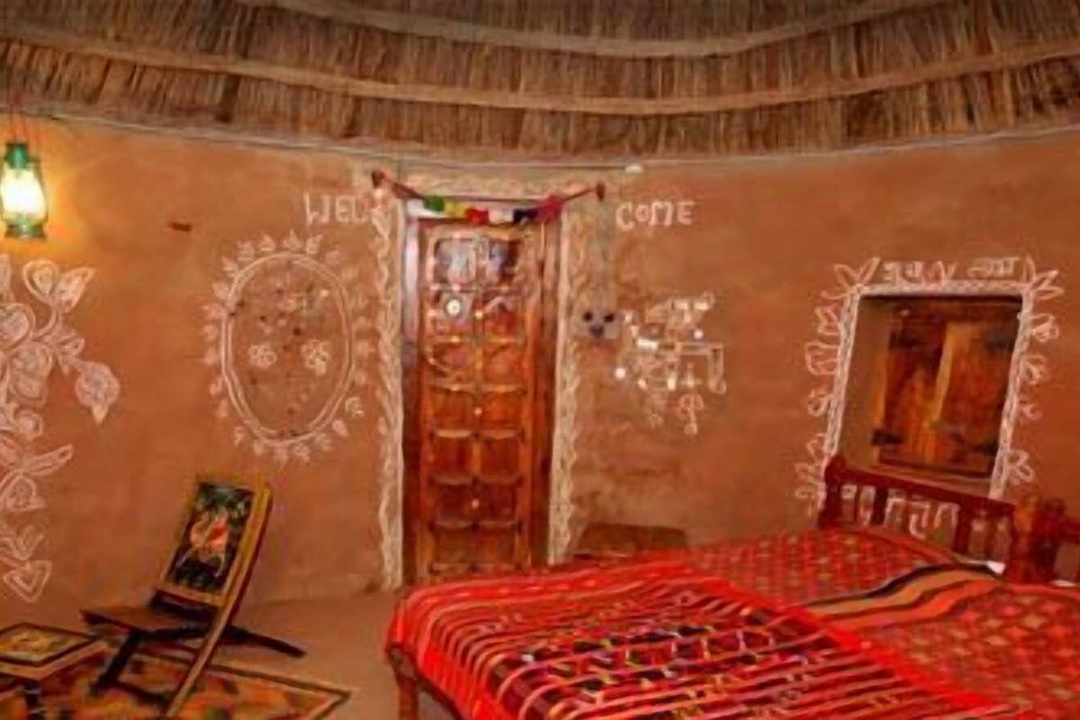 Une chambre traditionnelle avec un plafond de chaume, un lit recouvert d’une courtepointe à motifs rouges, une petite table avec une chaise et un mur orné de motifs décoratifs.