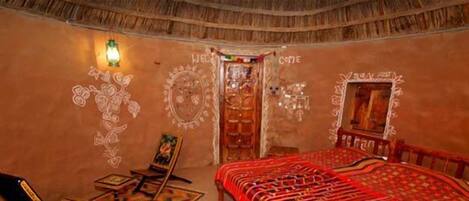 Una habitación tradicional con techo de paja, una cama con colcha estampada roja, una mesita con silla y una pared con motivos decorativos.