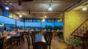 An outdoor dining area with a long table and chairs, surrounded by potted plants and a view of the cityscape at dusk.