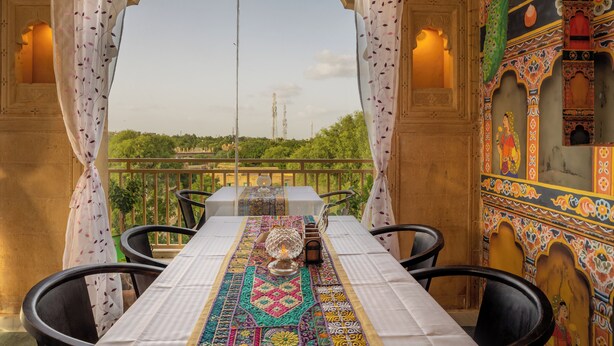 Zona pranzo con tavolo apparecchiato per due, caratterizzata da una tovaglia decorata e un centrotavola con candela. La stanza presenta pareti elaborate e un balcone con vista su spazi verdi e edifici in lontananza.