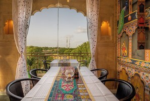 A dining area with a table set for two, featuring a patterned tablecloth and a candle centerpiece. The room has ornate walls and a balcony with a view of greenery and distant structures. - Hotel Meri Haveli (Jaisalmer)