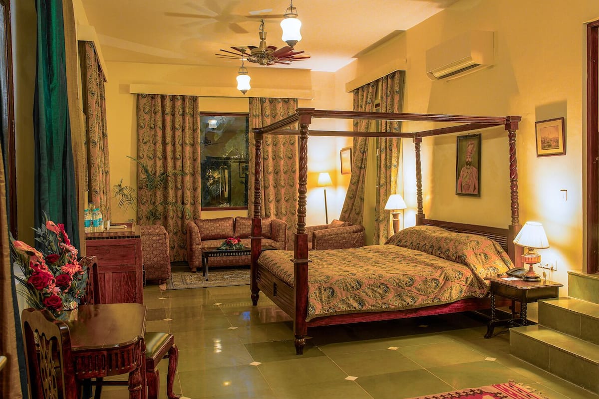 A four-poster bed with a floral bedspread, a sitting area with a sofa and armchairs, a desk with a lamp, and a large window with curtains.