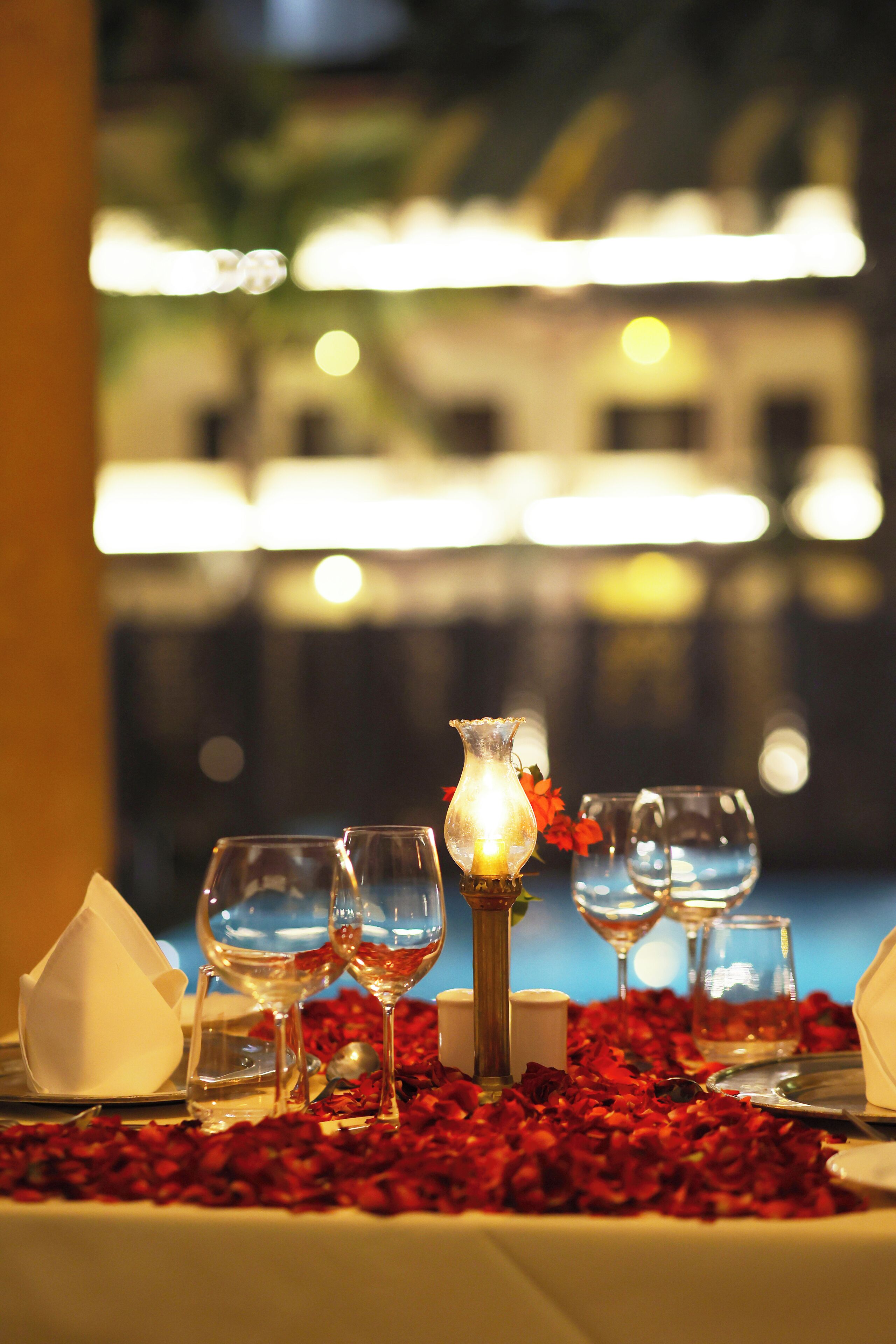 Una mesa dispuesta con copas de vino, una vela y pétalos de rosa, con un paisaje urbano borroso al fondo.