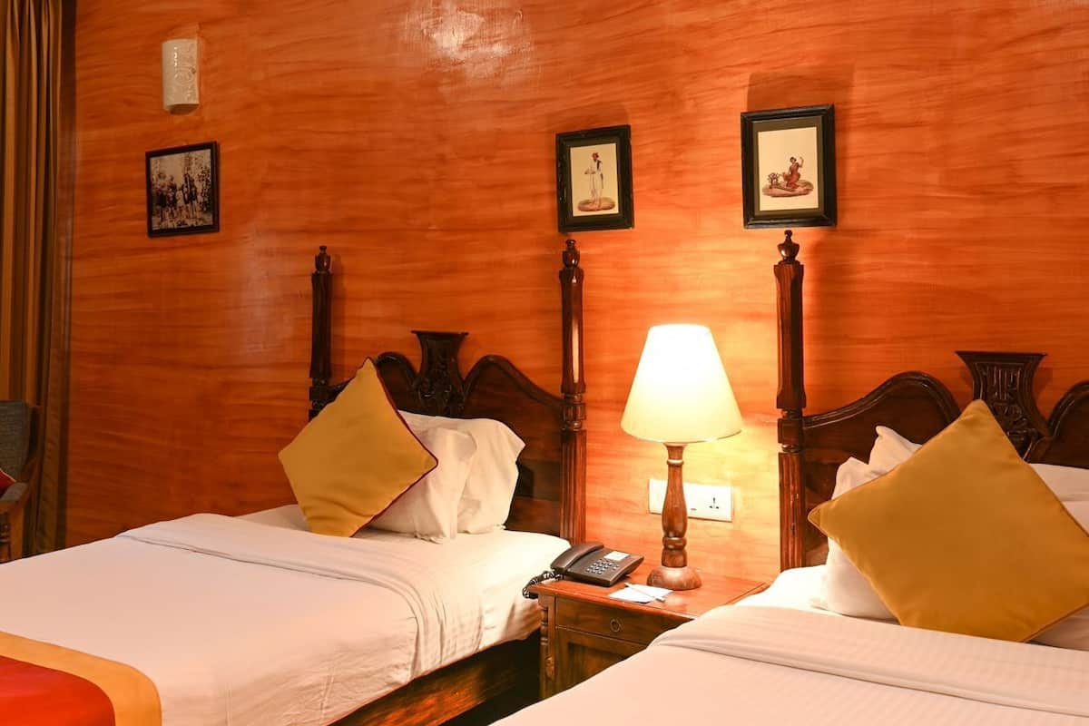 A hotel room with two beds, wooden headboards, a nightstand with a lamp, and framed pictures on the wall.