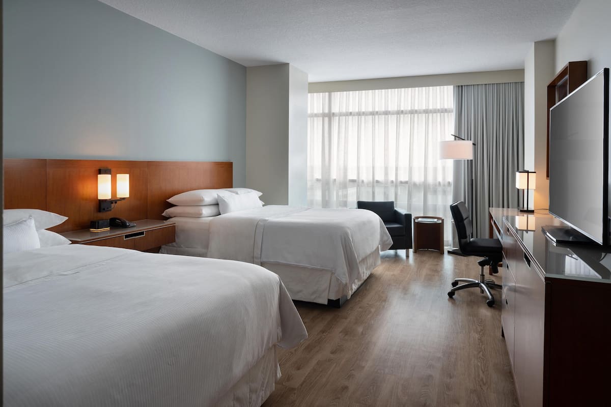 A hotel room with two beds, a desk, a chair, a TV, and a large window with curtains.