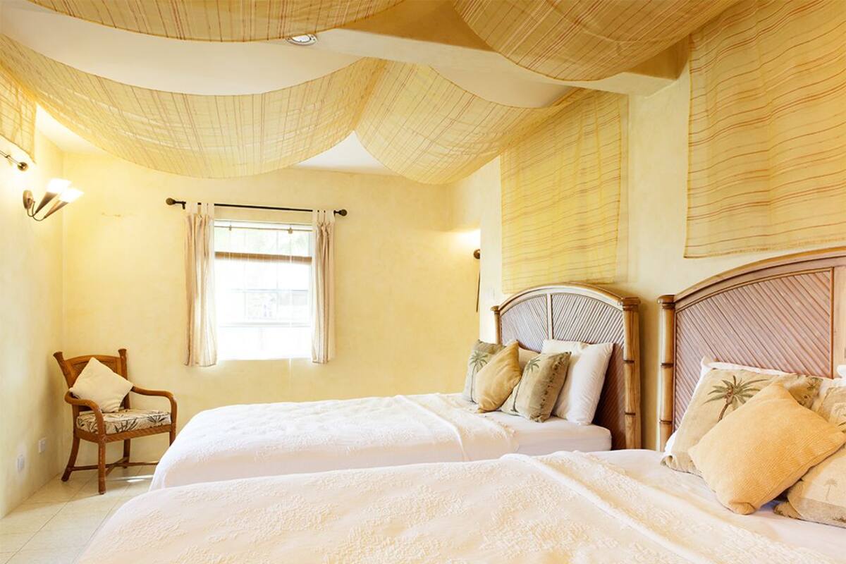Ein Zimmer mit zwei Betten, einem Sessel, einem Fenster mit Vorhängen und einer ansprechend gestalteten Decke.
