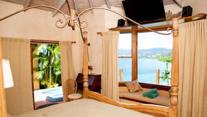 Un dormitorio con una cama grande, un asiento junto a la ventana con vistas al mar y un televisor sobre un mueble.
