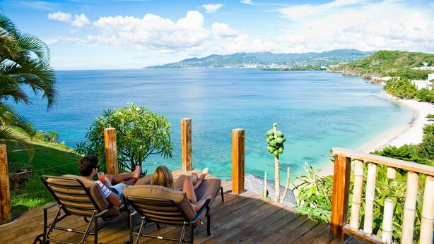 Una terraza de madera con vista a una playa, agua azul cristalina y montañas a lo lejos.