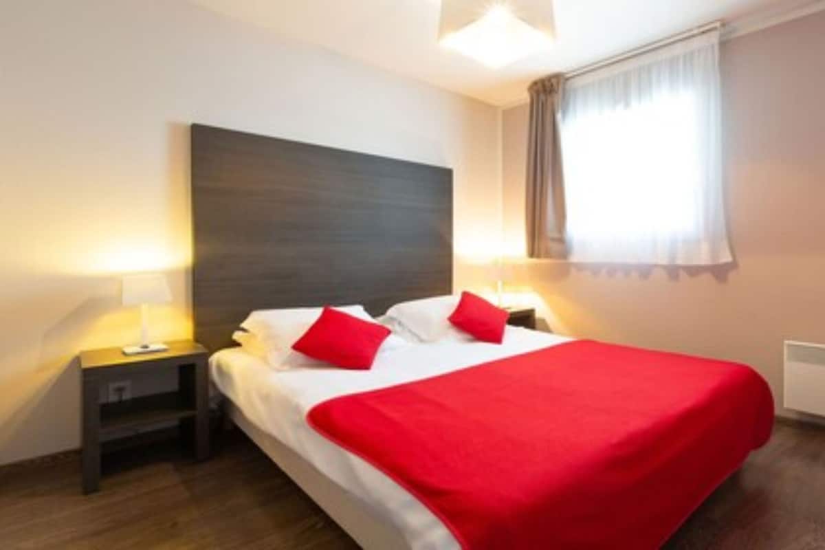 Ein Hotelzimmer mit einem großen Bett, zwei Nachttischen, einem Fenster mit Vorhängen und einer roten Tagesdecke.