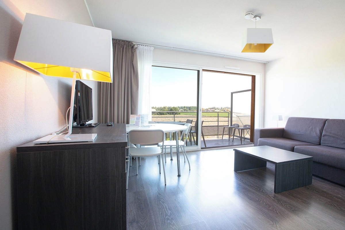 A modern hotel room with a desk, a grey sofa, a coffee table, and a dining area.