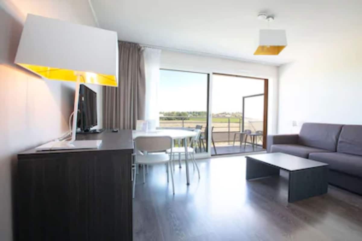 Ein modernes Hotelzimmer mit Schreibtisch, grauem Sofa, Esstisch mit Stühlen und einer großen Glastür zum Balkon.