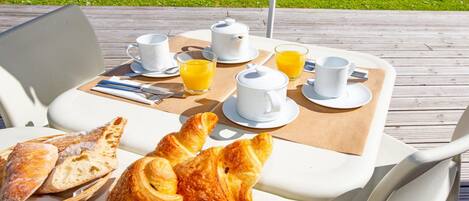 Una mesa exterior con desayuno que incluye cruasanes, pan, café y zumo de naranja.