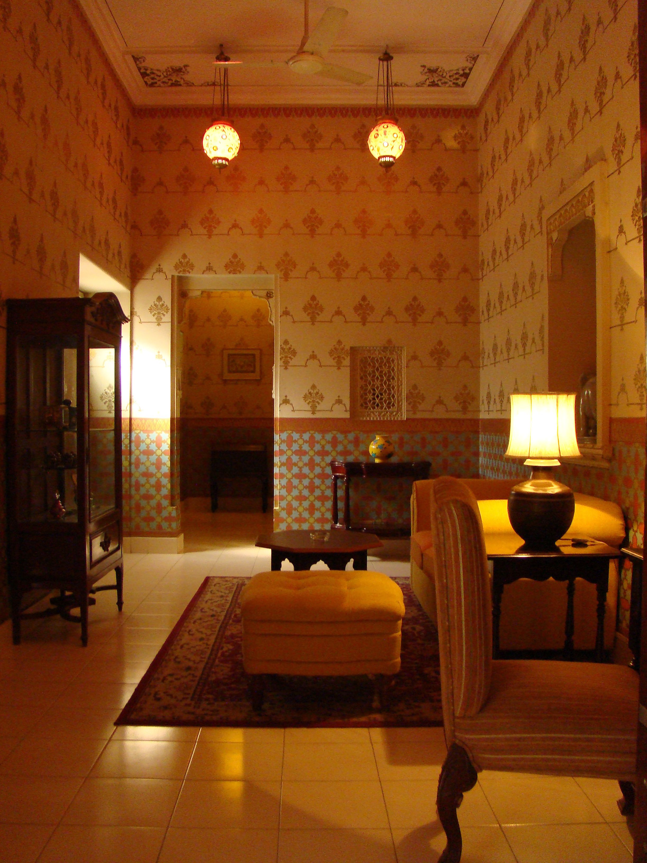 Ein Zimmer mit gemustertem Wandpapier, einem Holzschrank, einer Couch, einem Tisch mit Lampe und einem dekorativen Spiegel.