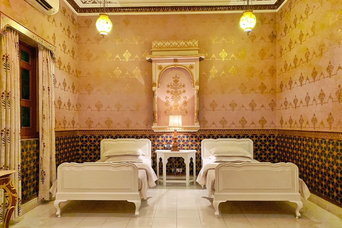 A room with two beds, a patterned wall, a decorative arch, and a small table with a lamp.