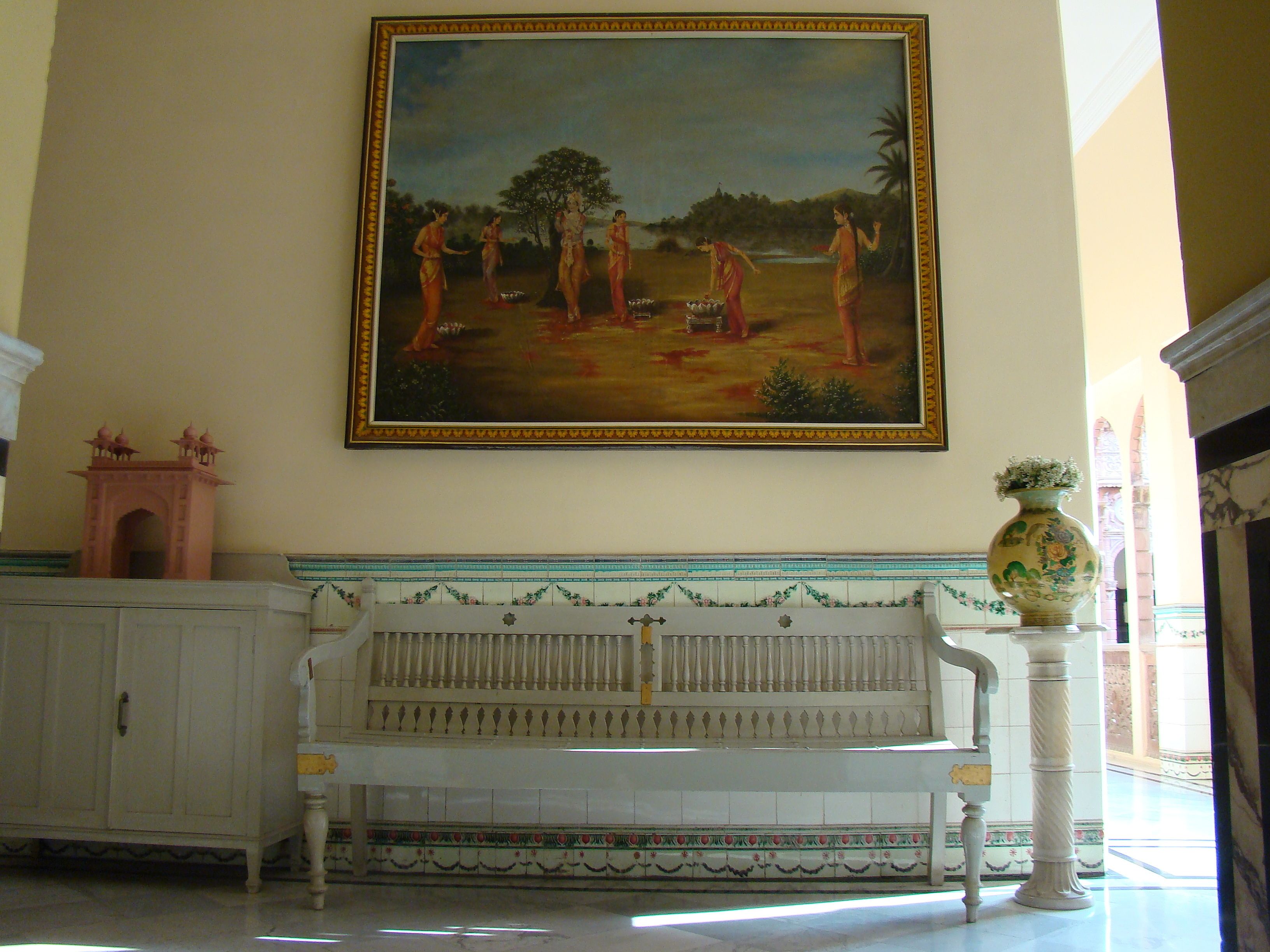 Una habitación con un banco blanco, un cuadro grande enmarcado en la pared y un jarrón decorativo sobre un pedestal.