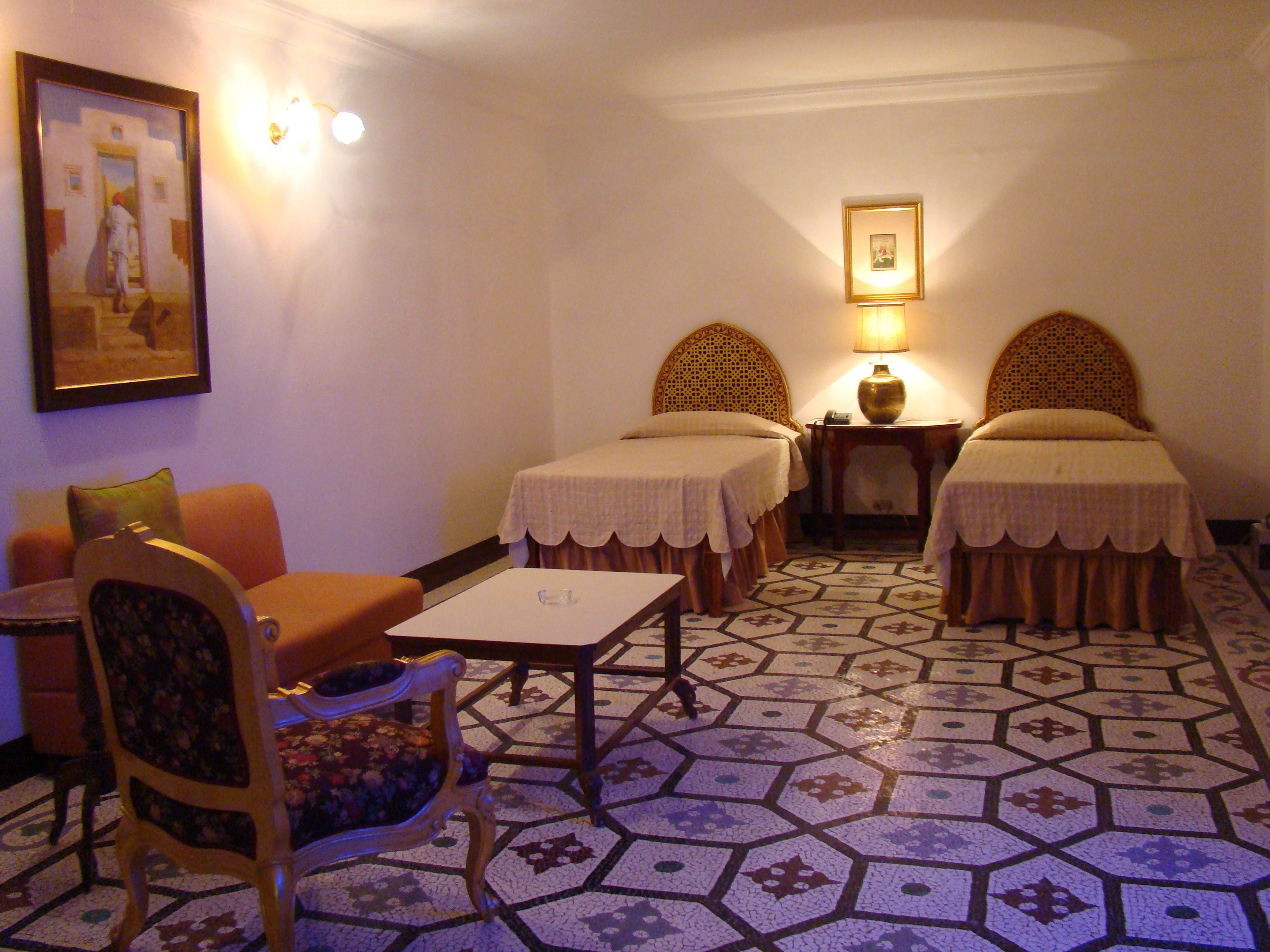 A hotel room with two single beds, a small table, a chair, a sofa, and a painting on the wall.