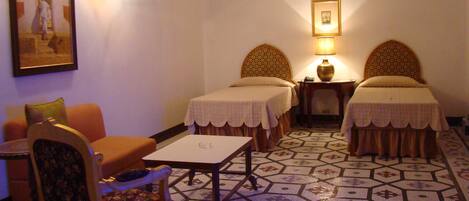 A hotel room with two single beds, a small table, a chair, a sofa, and a painting on the wall.
