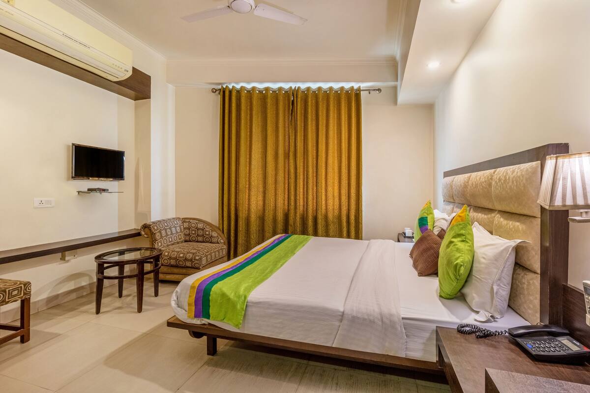 Una habitación de hotel con una cama, televisión montada en la pared, una silla, una mesita pequeña y un escritorio con teléfono.