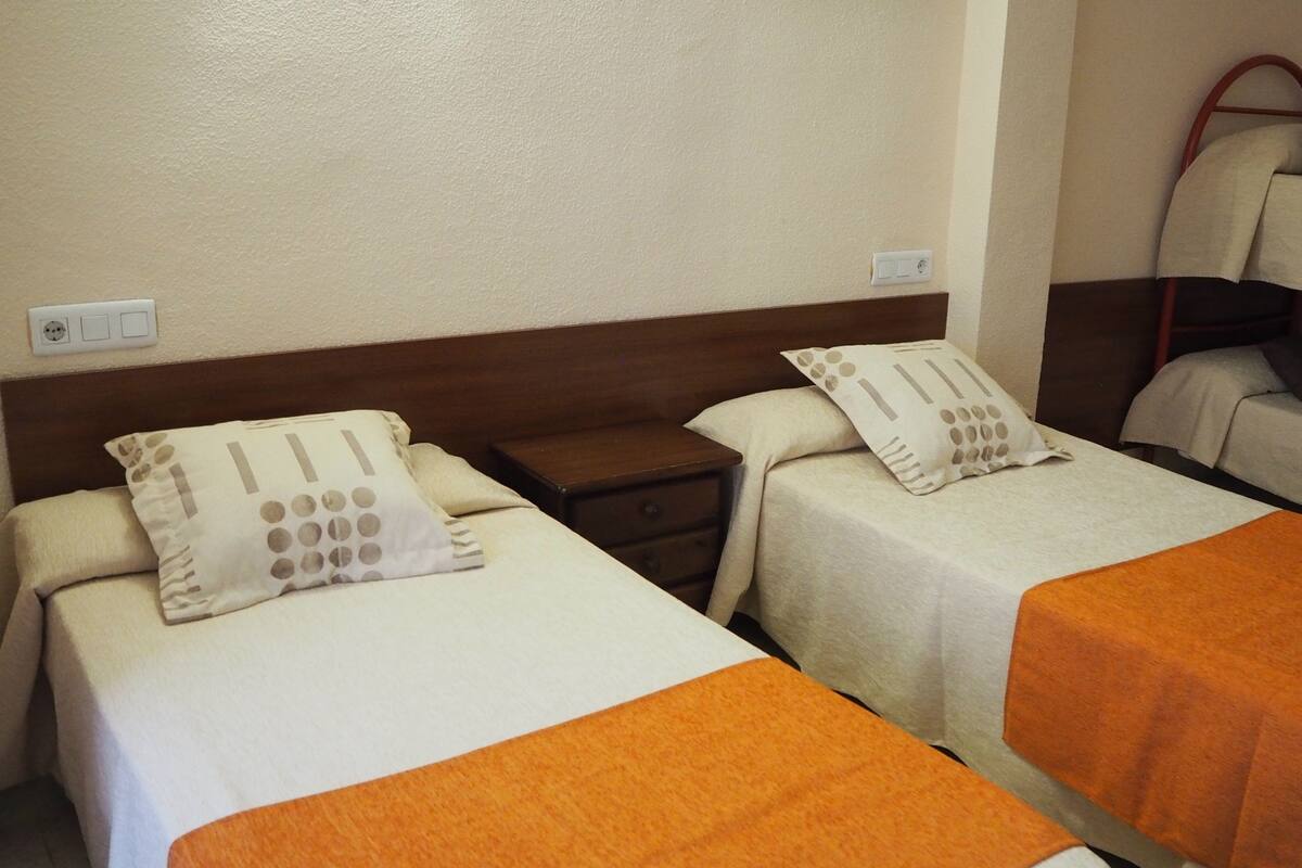 Two single beds with orange bedspreads, a wooden nightstand, and a bunk bed in the background.