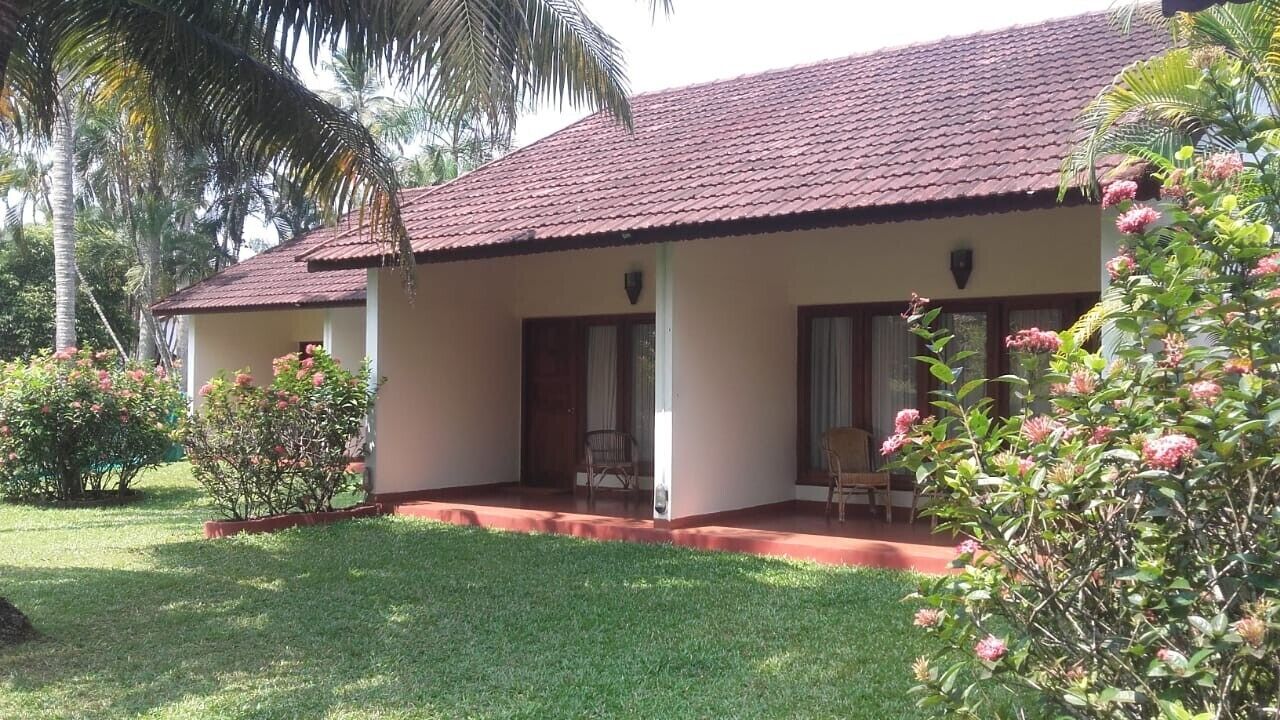 A two-story building with a red-tiled roof and a covered porch. There are two visible rooms with windows and doors. The building is surrounded by well-maintained grass and flowering plants.