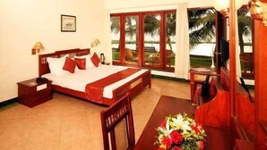 Un dormitorio con cama, mesita de noche, escritorio con silla, lámpara y ventana con vista a palmeras.