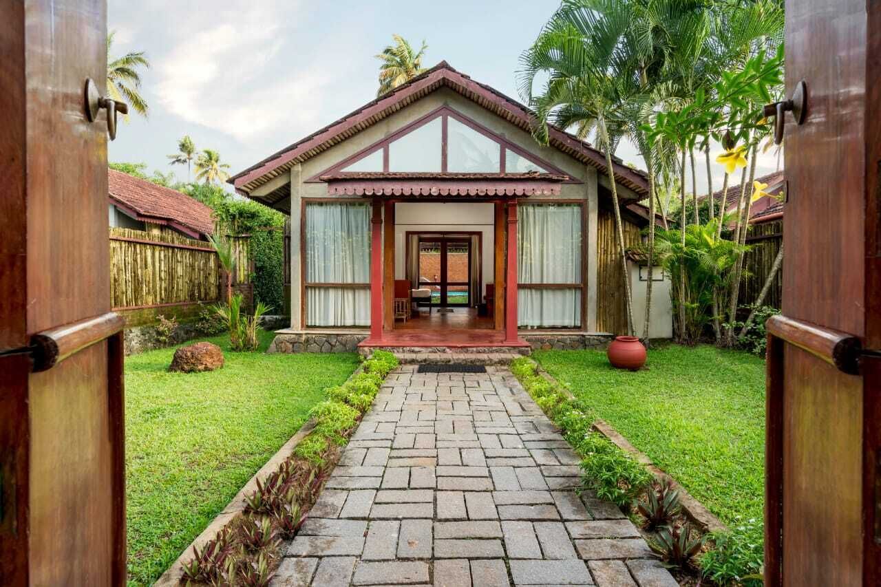 A bungalow with a tiled pathway leading to the entrance, surrounded by lush greenery and a wooden fence.