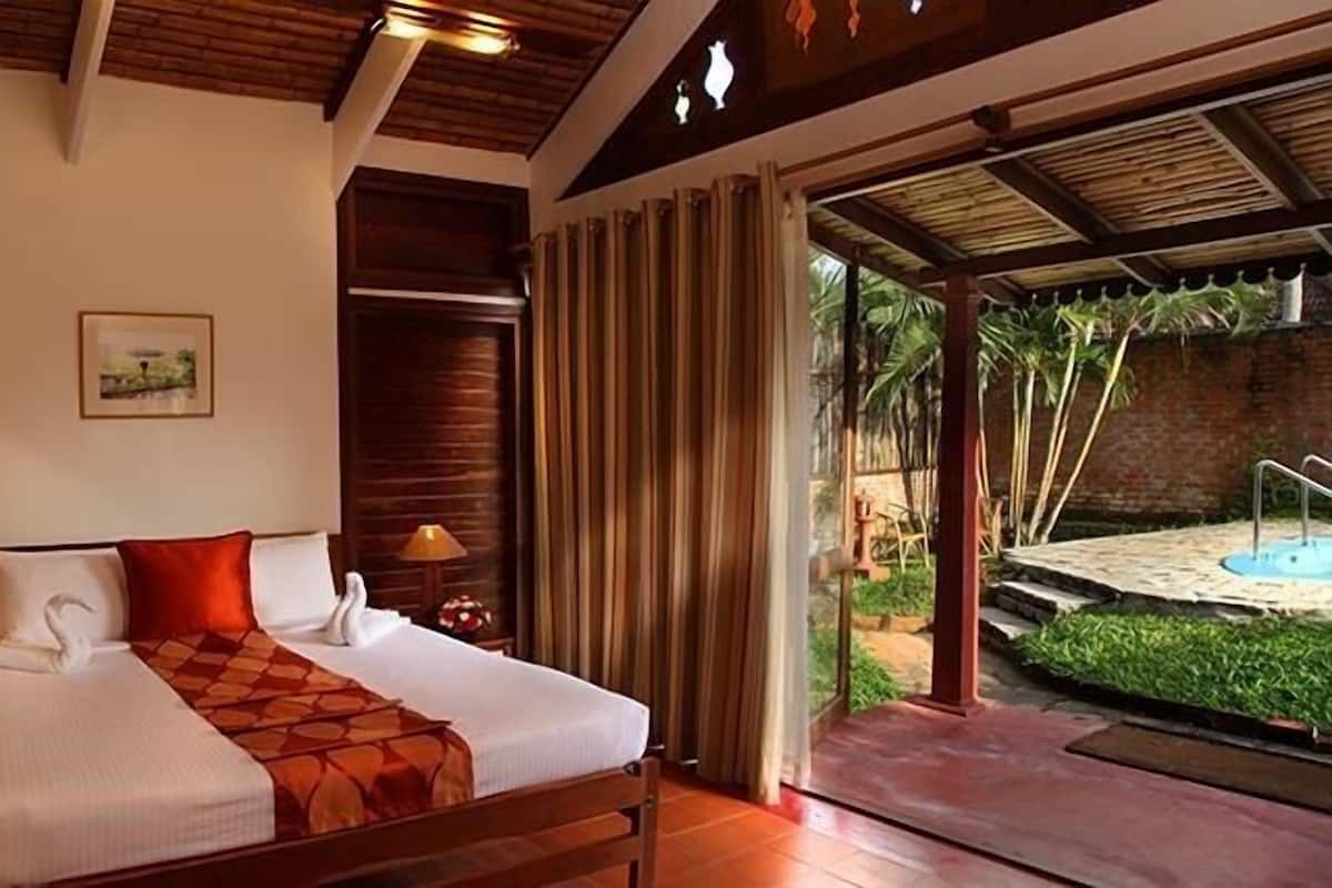 A hotel room with a bed, wooden headboard, two bedside tables with lamps, a small table with chairs, and a view of a pool and garden through a glass door.