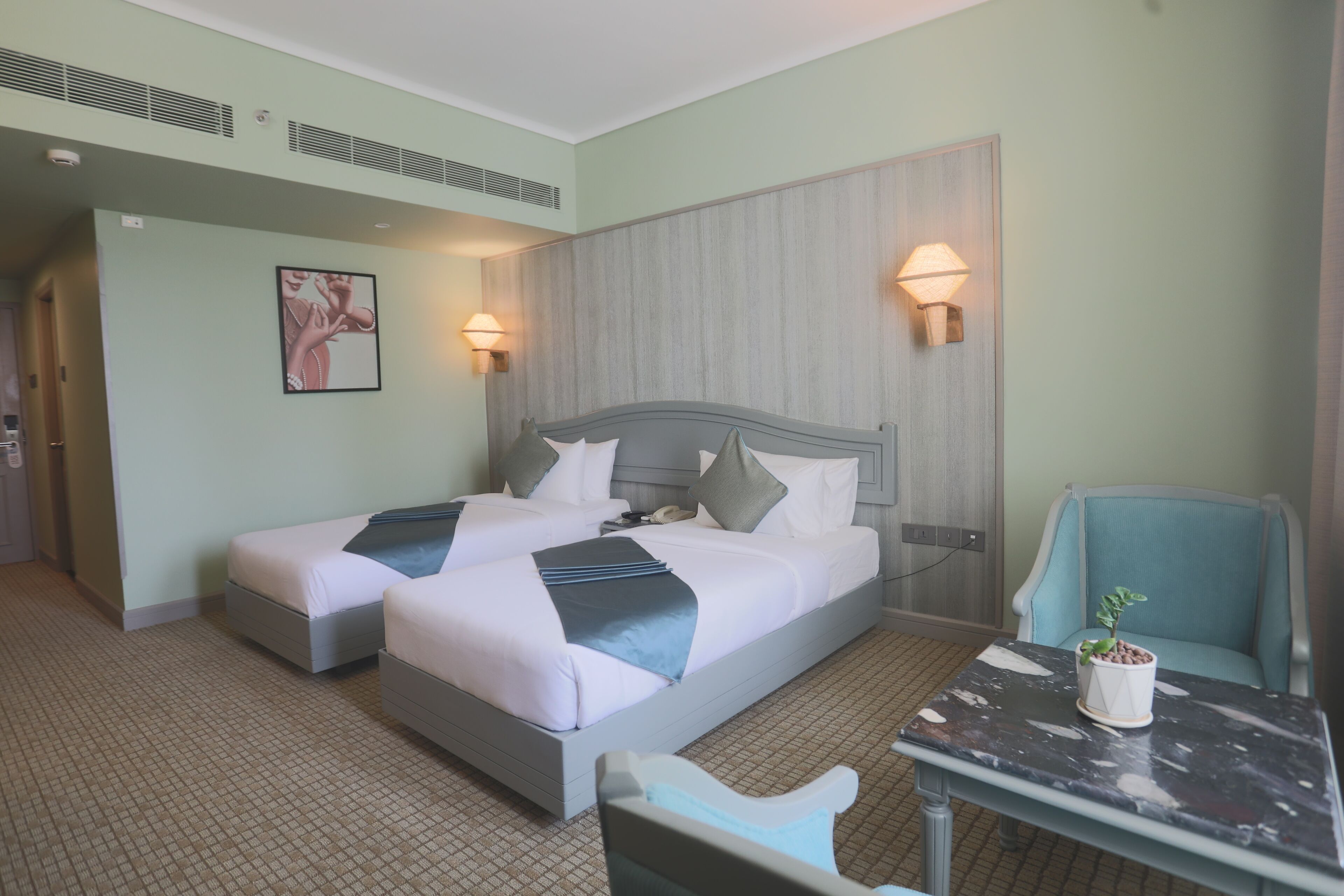 A hotel room with two beds, a small table, a chair, and a potted plant.