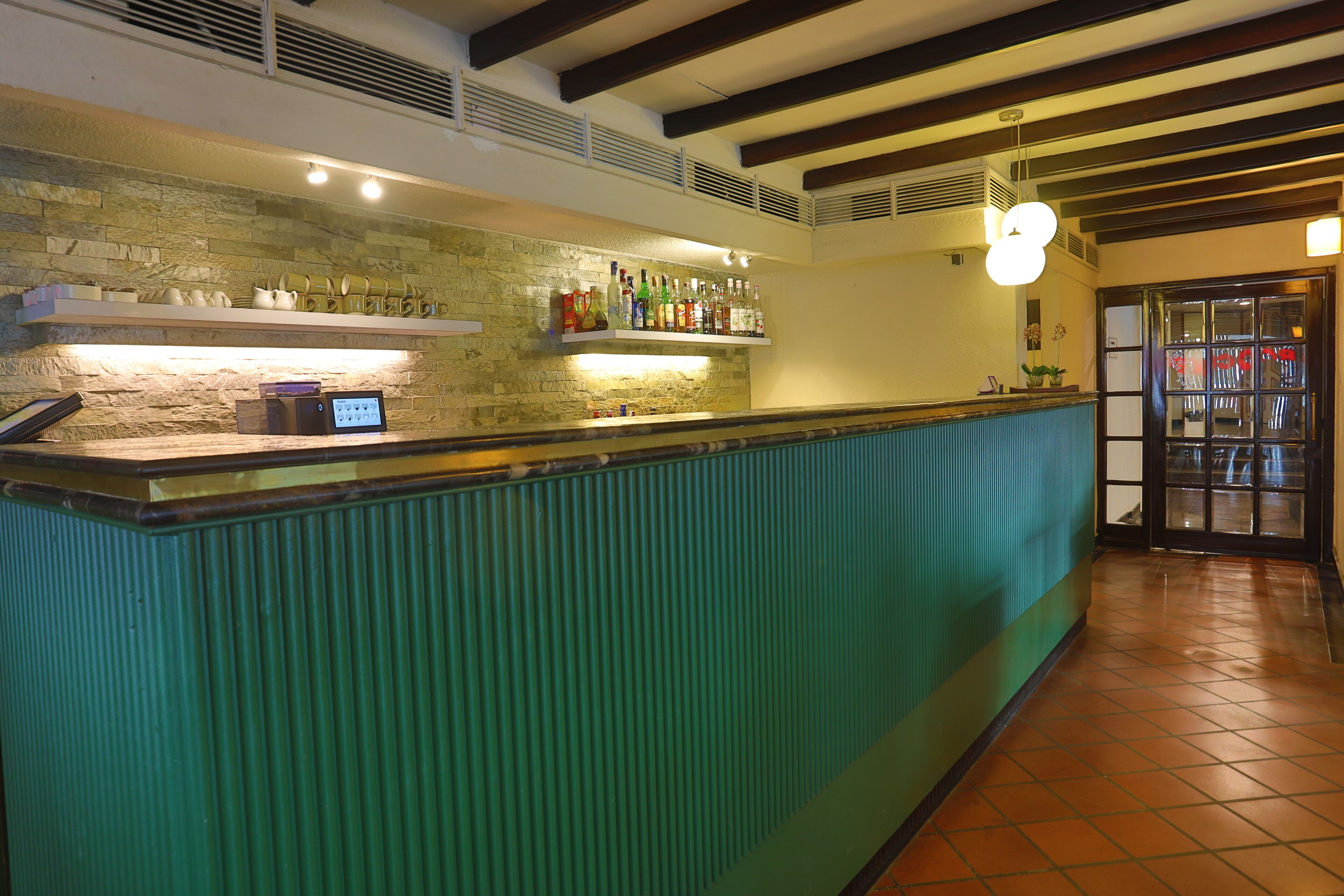 A bar area with a green counter, shelves with bottles, a stone wall, and a tiled floor.