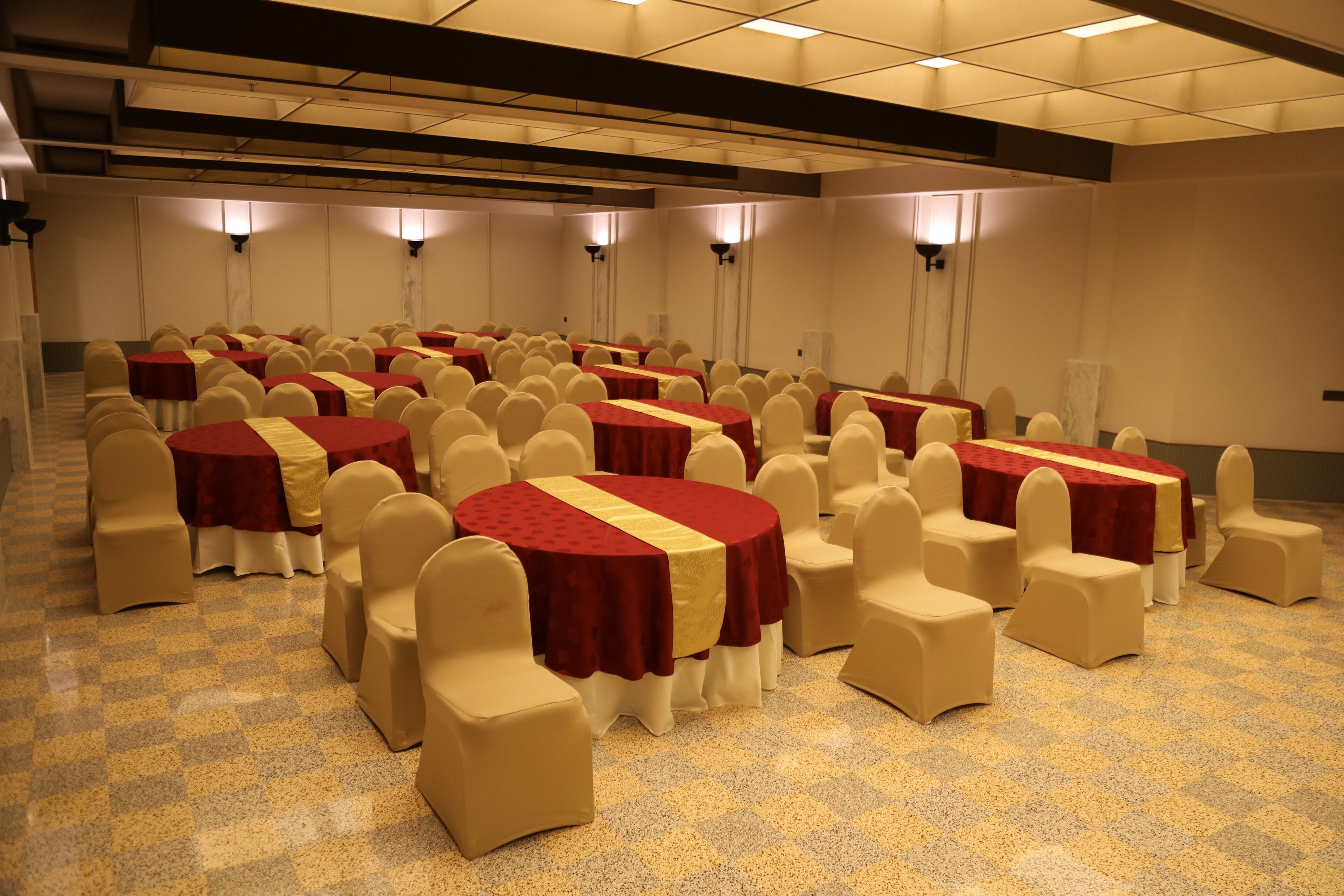 Un salón de conferencias con varias mesas redondas, cada una cubierta con un mantel rojo y amarillo, rodeado de sillas beige.