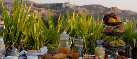 A breakfast setting with a table, a pitcher, a teapot, and a fruit display against a backdrop of greenery and a distant view of a hill.