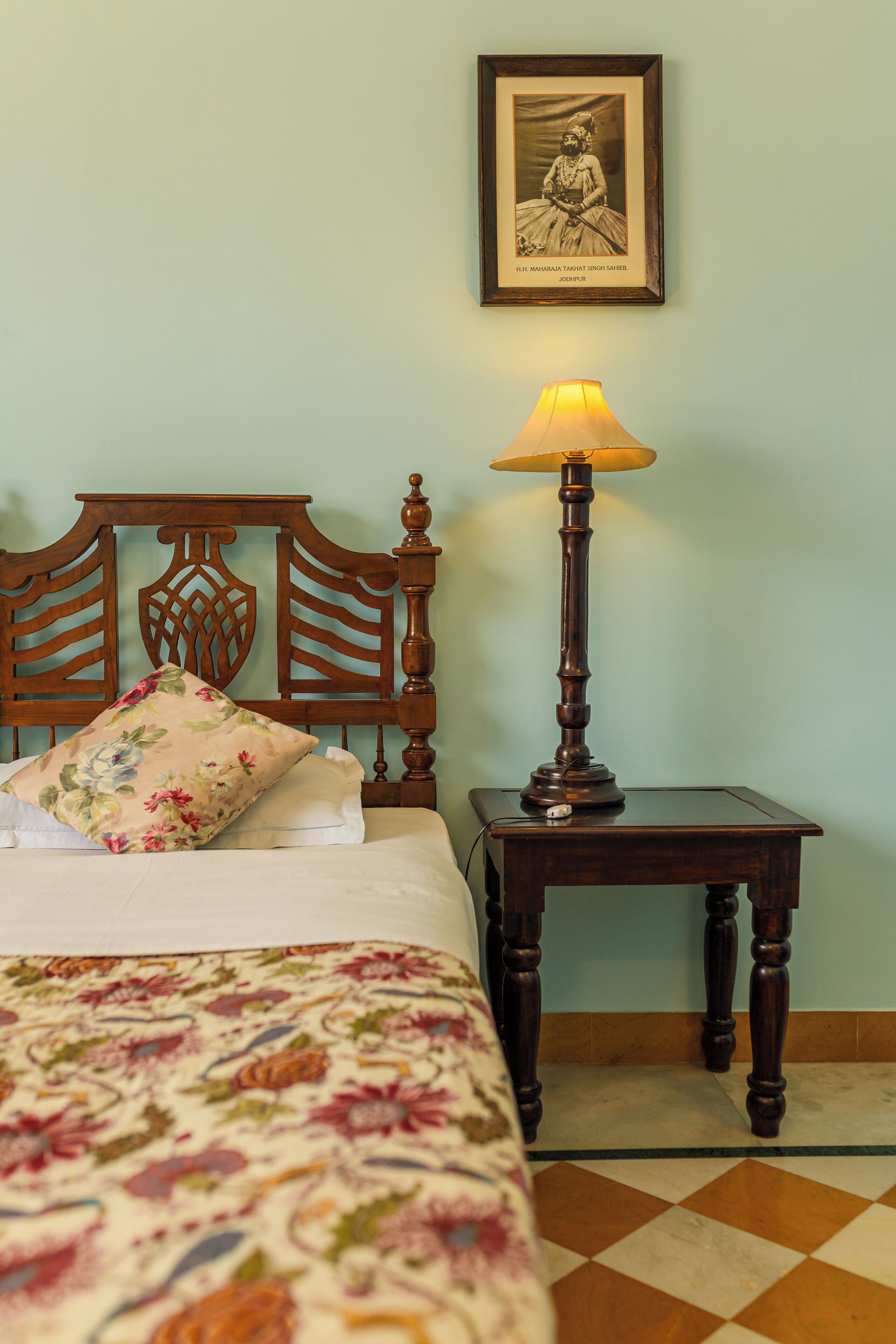 Una cama con colcha floral, cabecera de madera, mesita de noche con lámpara y un cuadro en la pared.