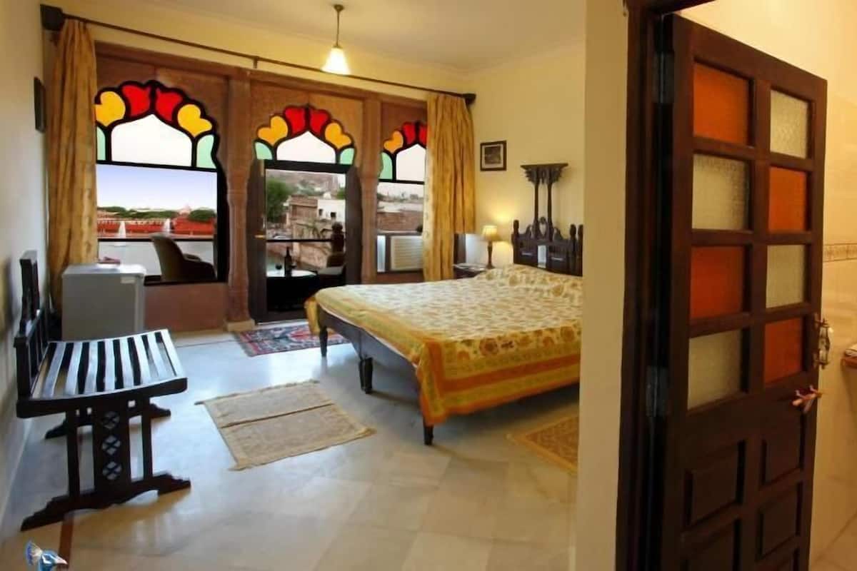 A hotel room with a bed, a desk, a chair, a window with stained glass, and a bathroom with a sink and mirror.