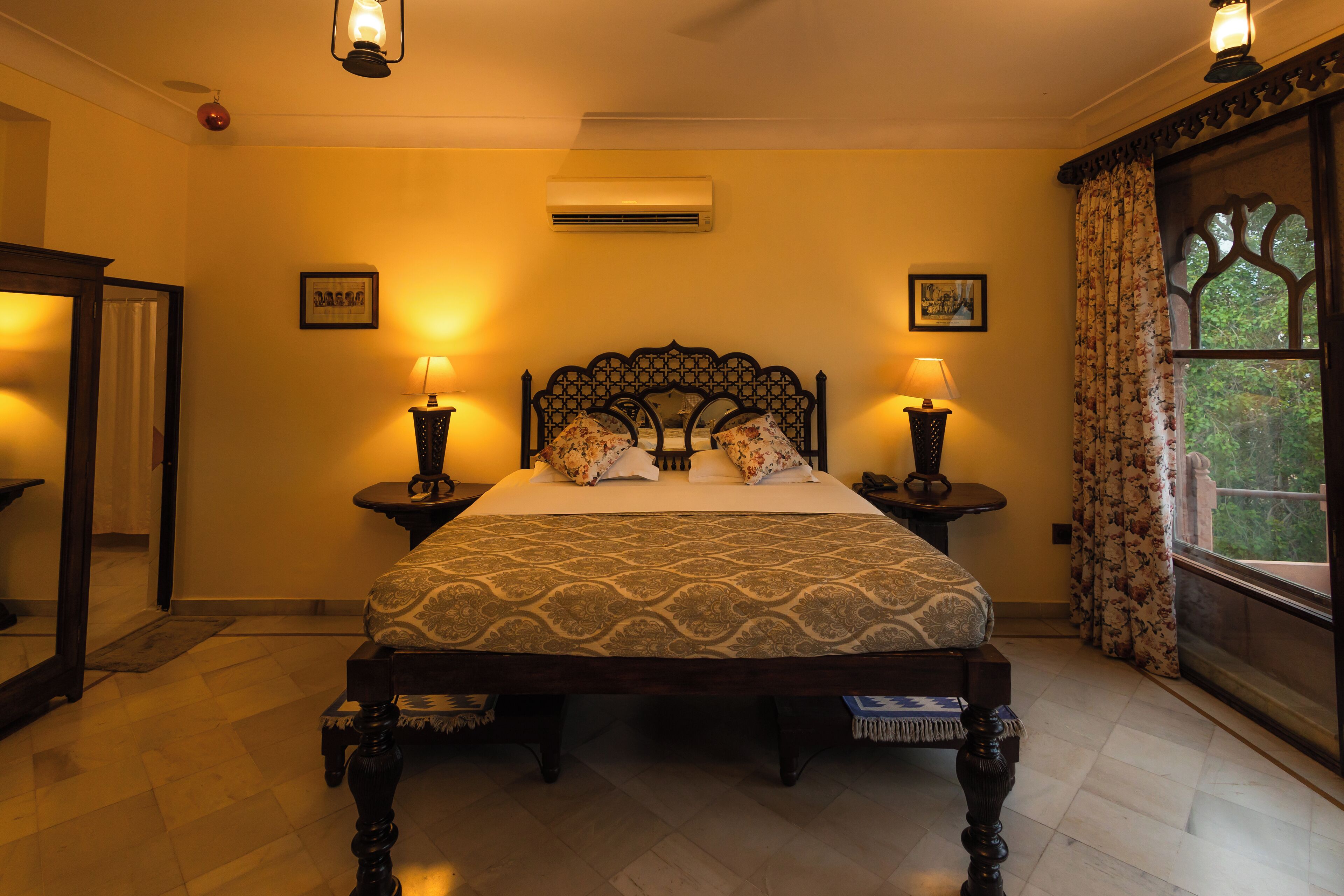 A neatly made bed with a patterned bedspread, two bedside lamps, and a window with curtains.