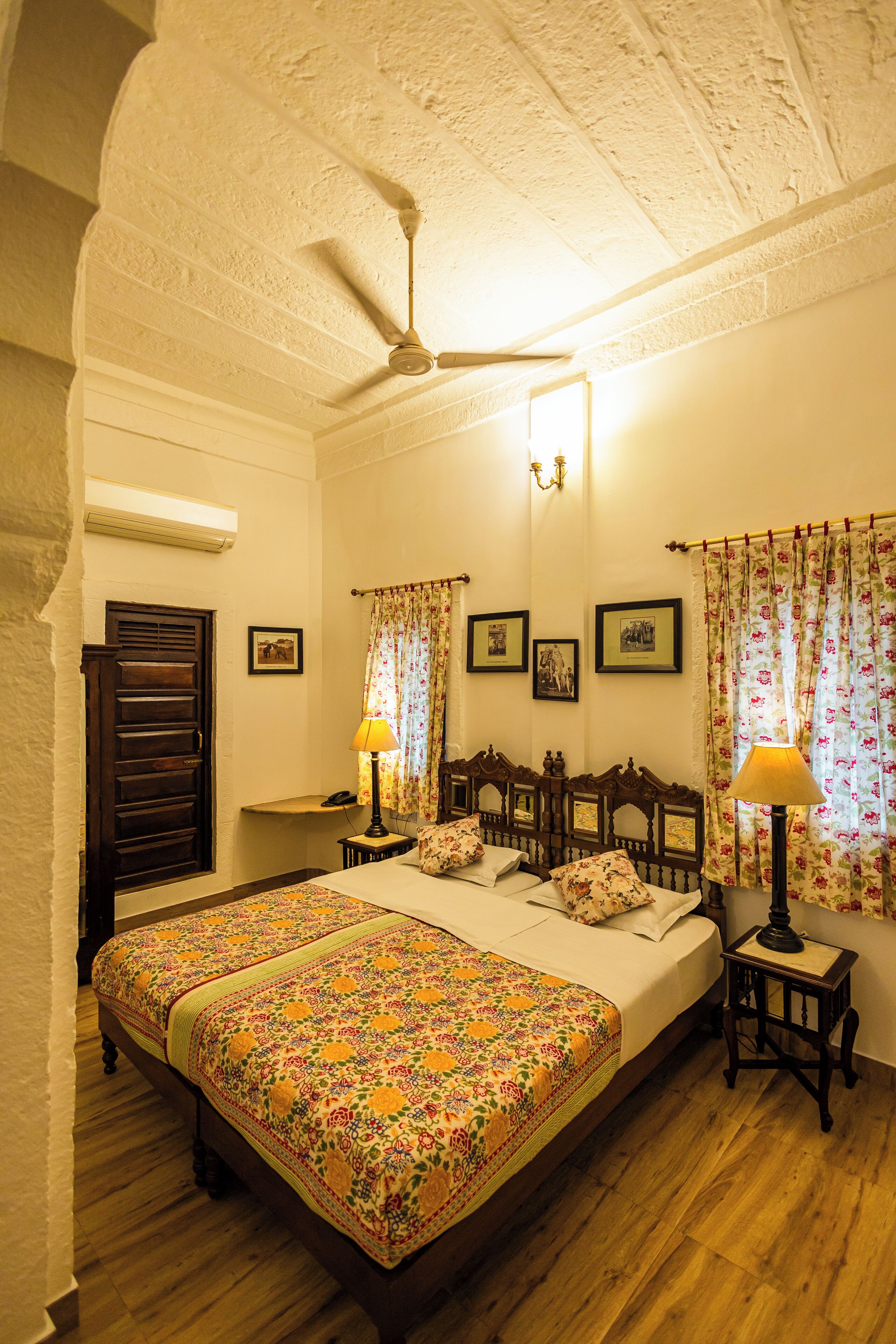 Un dormitorio con una cama grande, dos mesitas de noche con lámparas, un ventilador de techo y cortinas con motivos florales.