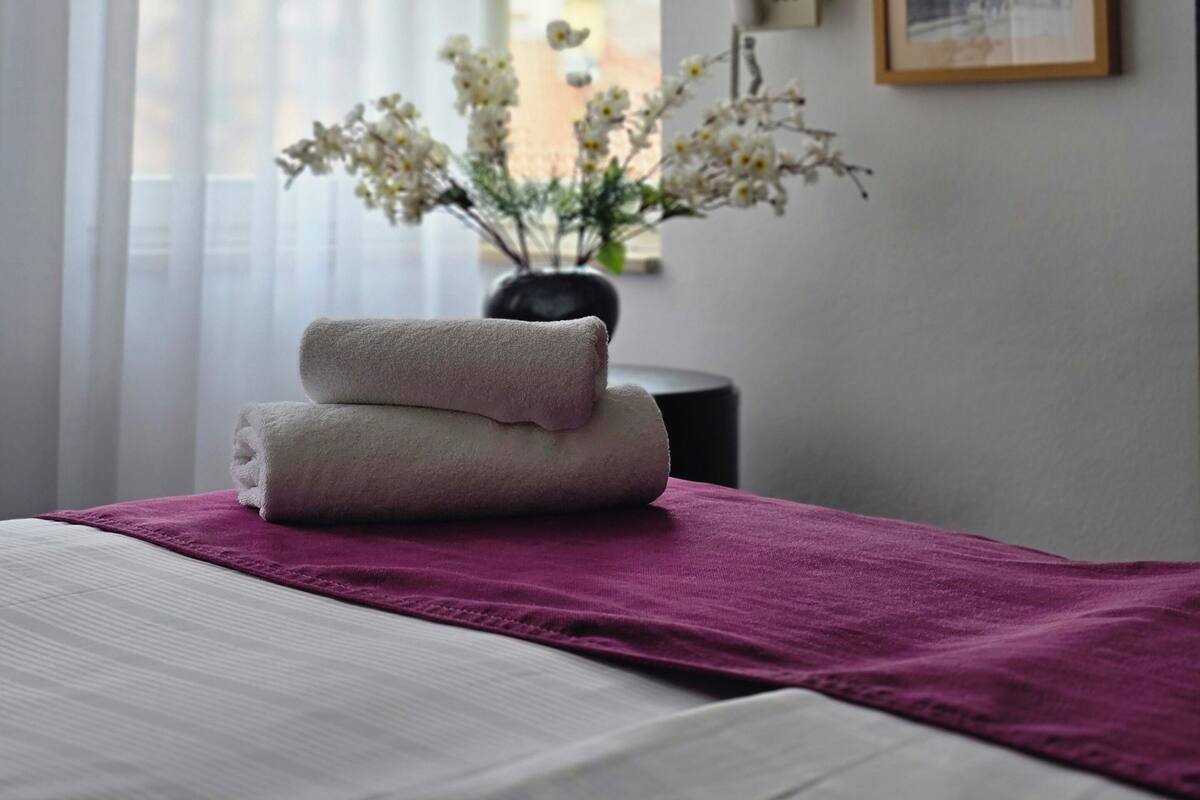 Habitación de hotel con una cama, una ventana con cortinas translúcidas, un jarrón con flores, un teléfono y un cuadro en la pared.