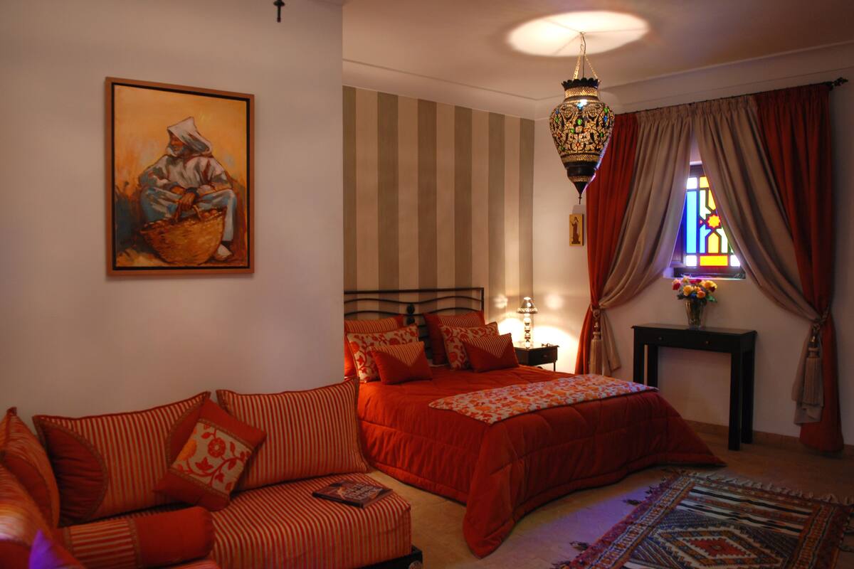 A bedroom with a bed, a sofa, a nightstand, a lamp, a painting, a window with curtains, and a hanging light fixture.