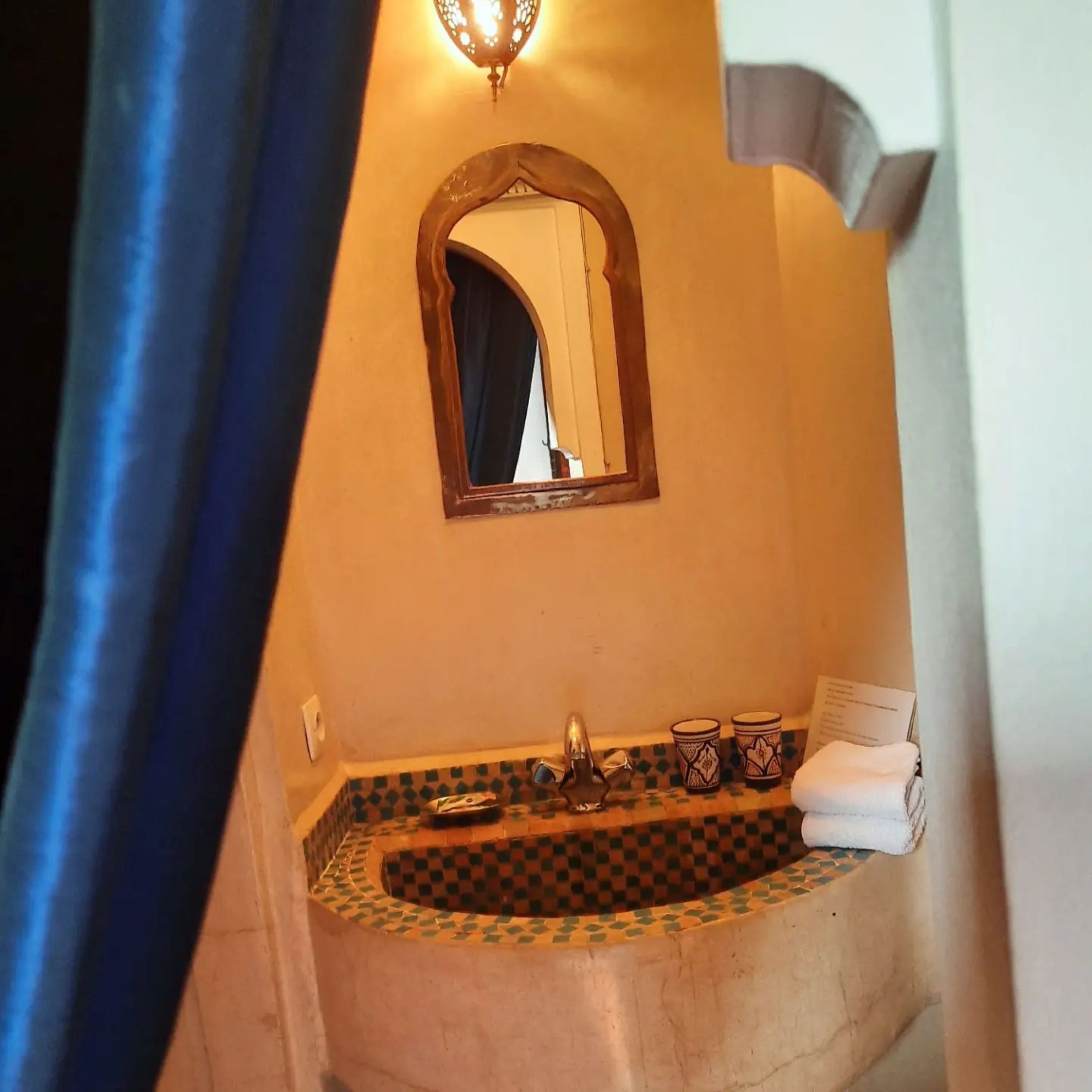 Une salle de bain avec un lavabo carrelé à motifs géométriques, un miroir doté d’un cadre en arc de cercle et un luminaire fixé au mur.