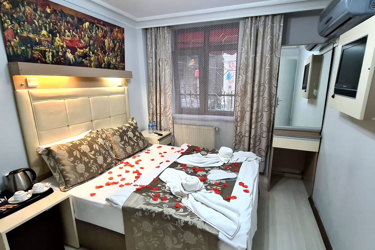 A hotel room with a bed, a nightstand, a painting, a window with curtains, and an air conditioning unit.