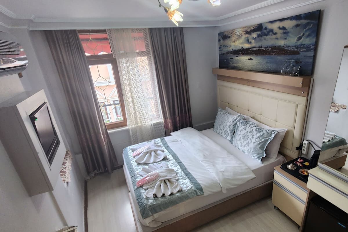 A hotel room with a bed, a nightstand, a painting, a window with curtains, and a view of the sea.