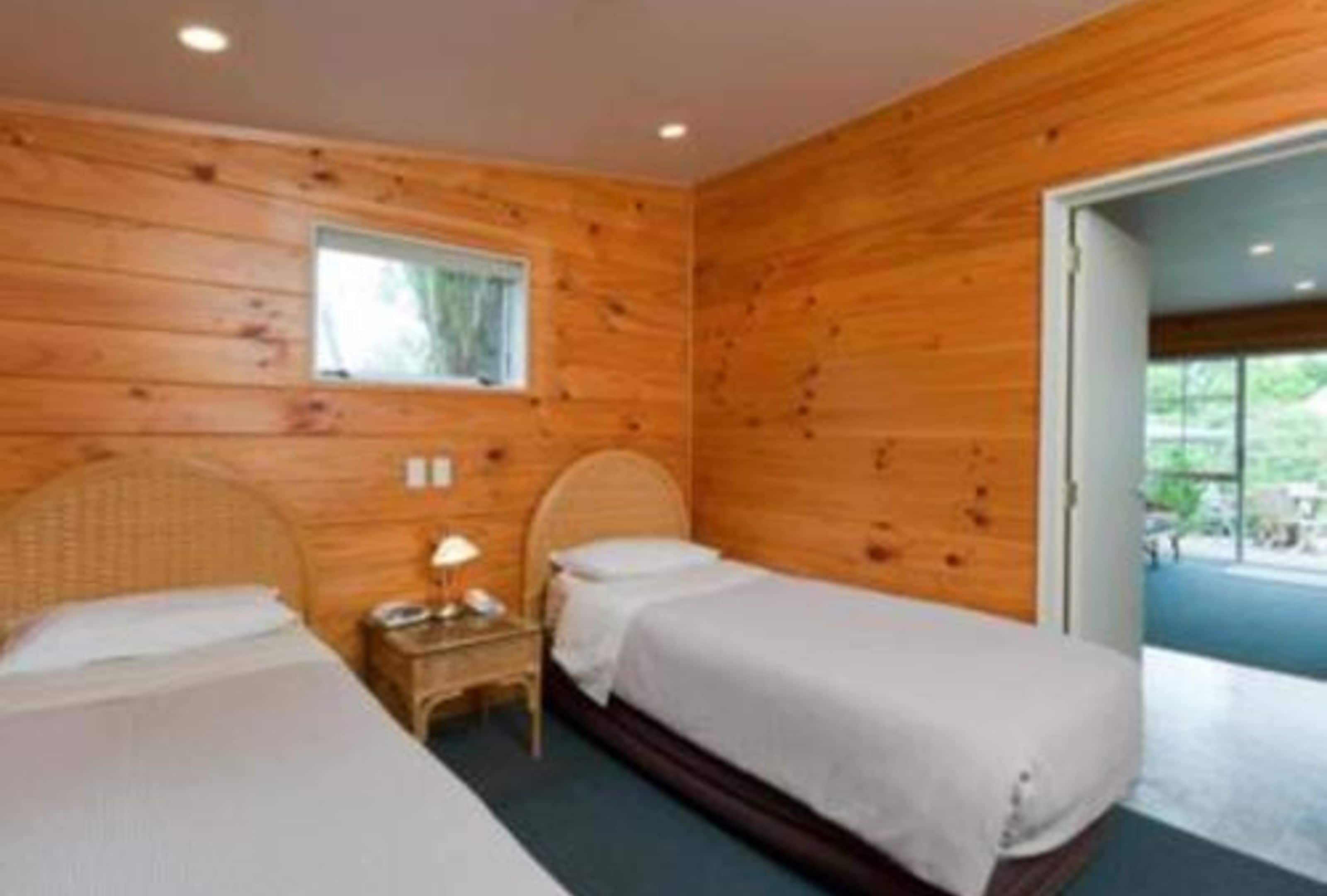 A room with two beds, wooden walls, a small table with a lamp, and a window.