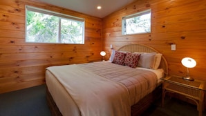 Habitación con paneles de madera, una cama, dos lámparas de noche y ventanas con vistas a árboles.