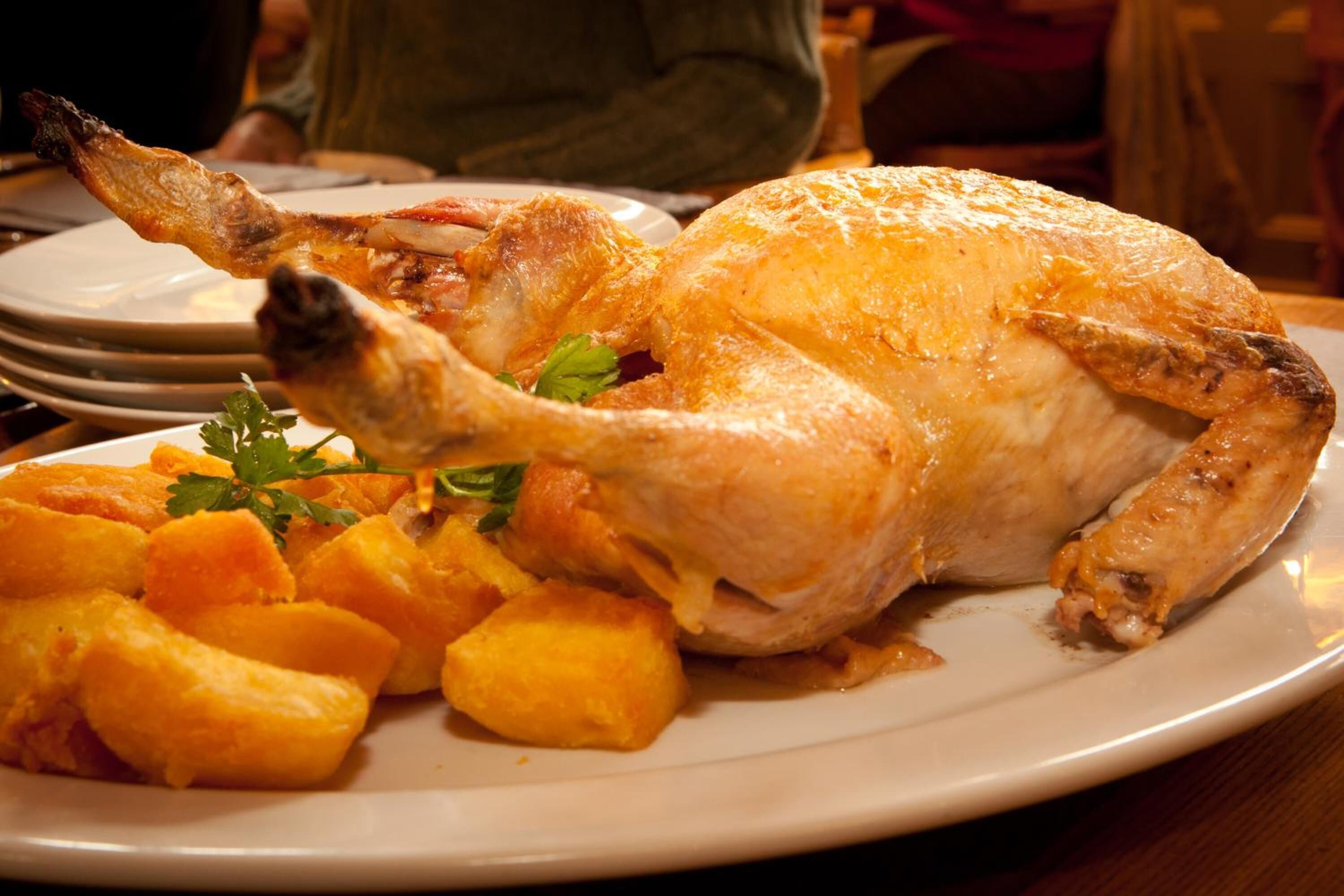 A roasted chicken with golden-brown skin, served on a white plate with golden-brown roasted potatoes and garnished with parsley.