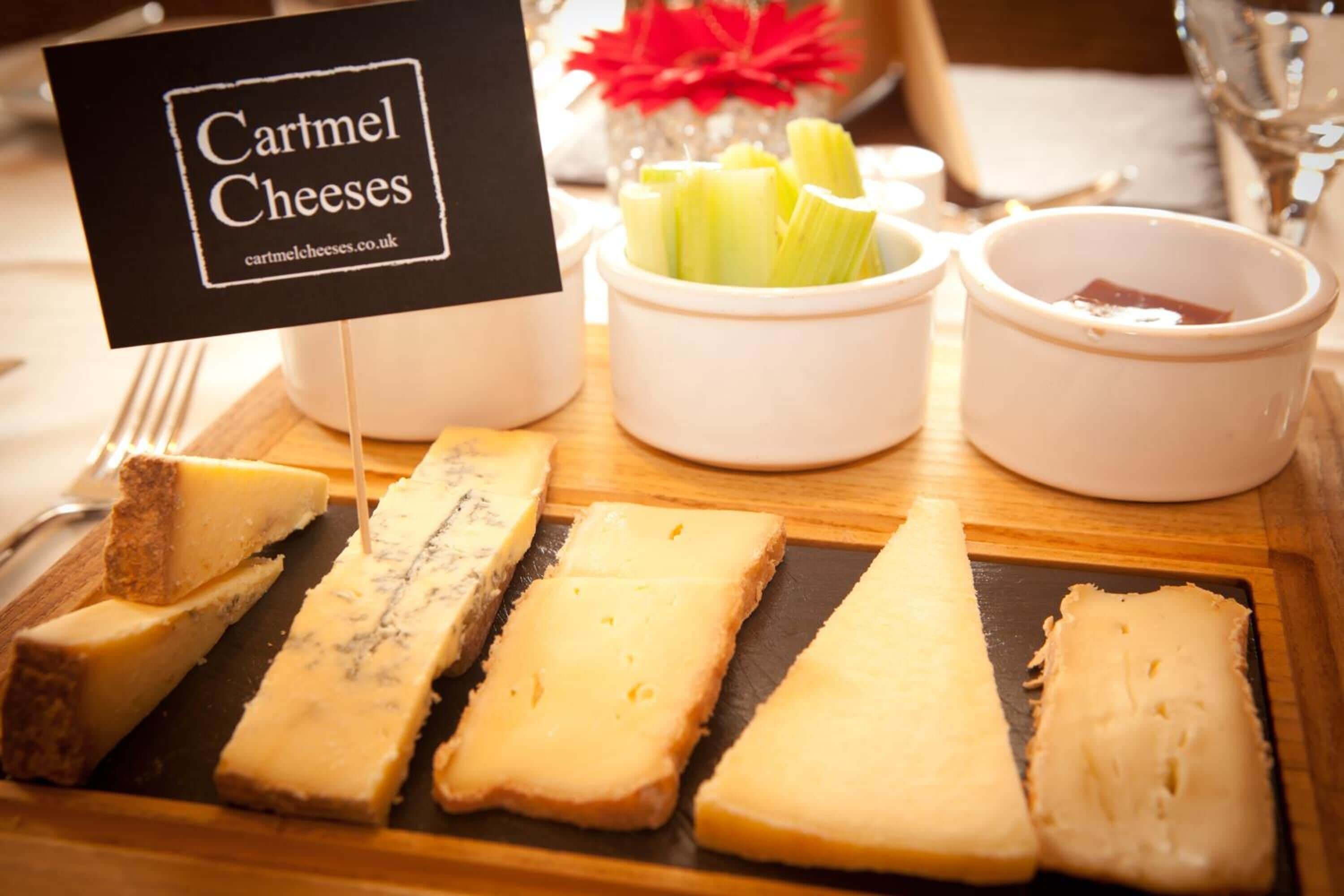 A cheese board with various types of cheese, celery, and a small sign that reads "Cartmel Cheeses."