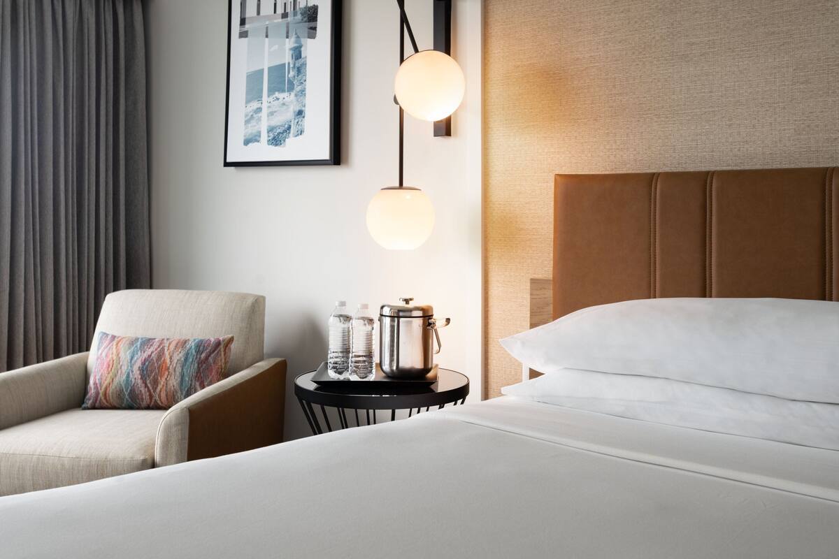 Une chambre d’hôtel avec un lit, une chaise, une petite table avec une bouilloire, des bouteilles d’eau et un tableau encadré au mur.