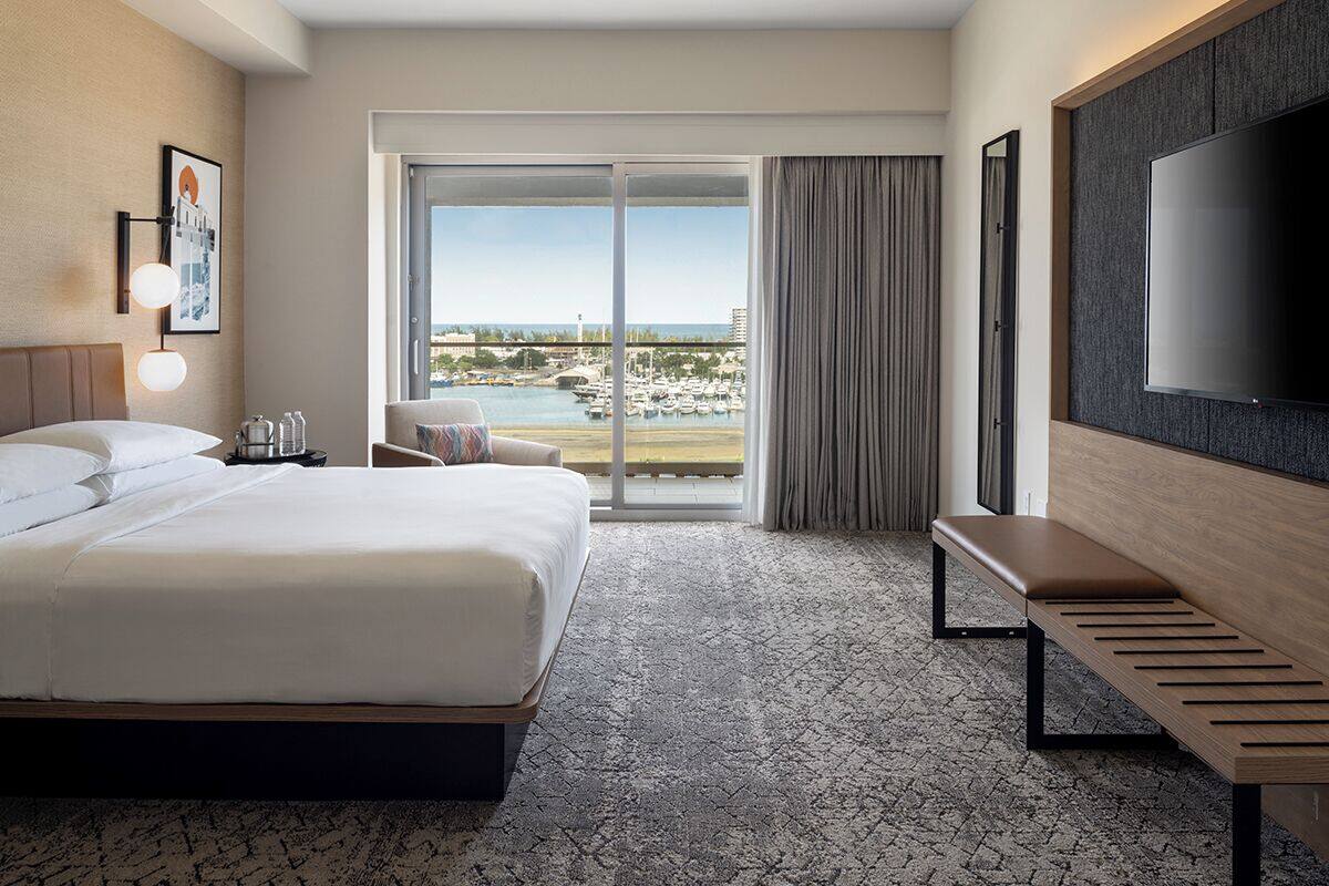 Une chambre d’hôtel dotée d’un grand lit, d’un téléviseur à écran plat, d’un banc et offrant une vue sur le port de plaisance à travers la porte vitrée.